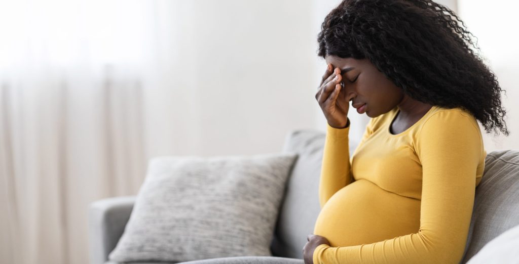 Young pregnant black woman suffering from headache or migraine, feeling sick, sitting on sofa at home, free space. Sad african american expecting lady touching her forehead and big tummy, home alone